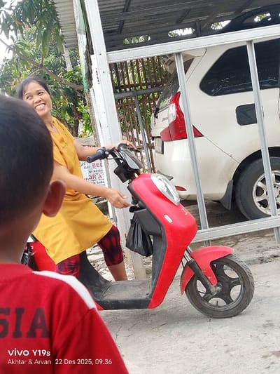 A woman in a yellow dress smiles while sitting on a red scooter. She is in front of a house with a car parked behind a metal fence. A child in a red shirt stands in the foreground with their back to the camera. Text at the bottom of the image reads "vivo Y19s, Akhtar & Arvan 22 Des 2025, 09.36." A sign is partially visible with the words "KBE ap Seda ie Seda".