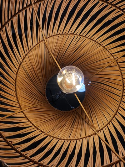 The image shows a close-up, downward-facing view of a light fixture with a woven rattan shade and a visible incandescent light bulb. The shade is circular with radiating spokes made of thin, dried plant material, creating a fan-like pattern. The light bulb is suspended in the center, emitting a warm glow that illuminates the surrounding weave. The background is dark, suggesting the light fixture is in an indoor setting, possibly at night. There are no people or other objects visible in the frame. The image is a high-angle shot, focusing on the texture and construction of the lampshade and the light source. The location of Maitland, United States of America, is not visually represented in the image.
