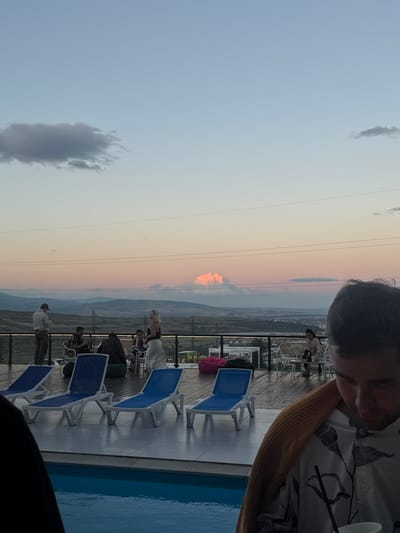 The image captures a twilight scene on a rooftop terrace in Mukhatskaro, Georgia. Several individuals are present, some seated at tables, others standing, engaged in conversation or observing the surroundings. A woman in a white skirt and black top is seen standing, while a man in a grey sweatshirt and cap stands apart. The terrace is furnished with blue lounge chairs, a pink beanbag chair, and white outdoor seating. Beyond the terrace, a city view unfolds with distant mountains, power lines, and a body of water. The sky displays a gradient of colors, transitioning from light blue to soft pink and orange, indicating the setting sun. The foreground features a portion of a swimming pool. A man, partially visible in the lower right, is draped with a tan blanket.