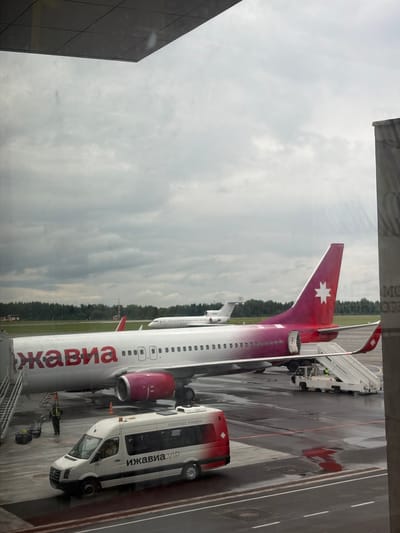 The image captures an airport scene, likely viewed from within a terminal building through a window, looking out onto the apron. The sky is overcast and cloudy, suggesting a daytime setting with no direct sunlight. The tarmac below appears wet in several areas, possibly from recent rain or cleaning.
The primary subject is a passenger aircraft, identifiable as belonging to the "ижавиа" (Izhavia) airline, based on the red Cyrillic text on its white fuselage. The plane features a distinctive red and magenta tail fin with a white star logo, and its engines are also magenta. The aircraft registration "RR-73062" is visible on the tail.
Adjacent to the plane, a white airport shuttle bus is parked, also bearing "ижавиа.VIP" and "Аэропорт Ижевск" (Izhevsk Airport) text on its side, confirming the location and airline. "Sprinter" is also visible on the front of the bus. Mobile passenger stairs are connected to the aircraft, indicating ongoing ground operations such as boarding or disembarkation. At least two airport ground crew members are visible near the aircraft and equipment, engaged in their duties.
In the background, a distant treeline separates the airport area from what appears to be a natural or less-developed landscape. The visible texts "Izhavia" and "Izhevsk Airport" strongly suggest the location is Izhevsk Airport, situated near Новая Казмаска, Russia.