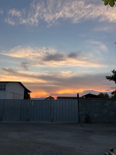 The image depicts a sunset over a partially constructed or industrial area in Dolni Dabnik, Bulgaria. The sky is filled with wispy clouds tinged with hues of orange, pink, and blue, indicating the sun is low on the horizon. In the foreground, a large, corrugated metal gate dominates the scene, with a dirt or gravel-covered ground extending towards it. To the left of the gate, a white building with a dark-tiled roof is visible. To the right, a wall constructed of cinder blocks stands, with a sparse plant growing near its base. Silhouetted structures, possibly roofs of other buildings or unfinished constructions, are visible behind the gate and the cinder block wall. The overall impression is one of a quiet, possibly undeveloped or industrial outskirts of the town at dusk. There are no people or animals visible in the image.