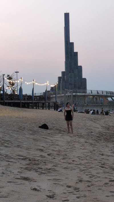 A woman in a black dress stands on a beach in Unknown, Vietnam, next to a tall, stepped, block-shaped building. The beach is sandy, and there are footprints in the sand. There is a black object (possibly a bag) near her, and some seaweed. A line of light bulbs is strung along a structure near the beach. A crowd of people is sitting on or near the building.
