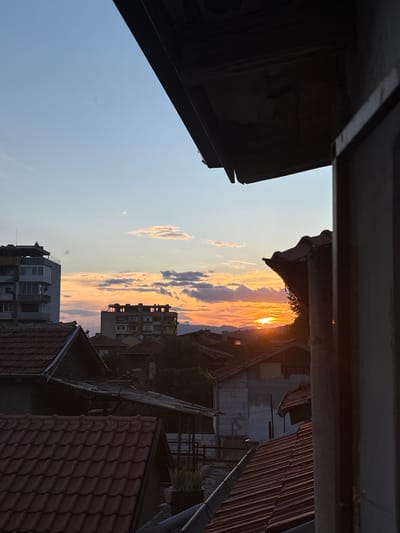 This is a vertical, eye-level photograph of a sunset over a residential area in Dupnitsa, Bulgaria. The sky is a vibrant mix of orange and blue, with scattered clouds tinged with the warm hues of the setting sun. The sun itself is partially obscured by clouds, creating a bright, glowing effect.
The foreground of the image is dominated by the dark shapes of overhanging structures, likely balconies or roofs, which frame the view of the town. Below these structures, a dense cluster of buildings with red-tiled roofs and varying architectural styles is visible. Some buildings appear to be older, while others are more modern, with multiple stories and balconies. Trees are interspersed among the buildings, adding a touch of greenery.
The time of day is clearly sunset, indicated by the dramatic colors of the sky and the low angle of the sun. The weather appears clear and calm. The location context of Dupnitsa, Bulgaria, is provided. No specific activity or event is evident, and no text is visible in the image. The overall mood is serene and picturesque, capturing the beauty of a sunset over a town.