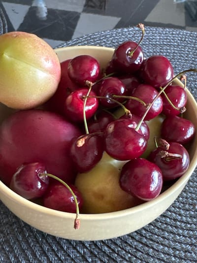 A close-up, top-down view shows a bowl filled with fresh fruit, including cherries and what appear to be plums or nectarines. The fruit is arranged in a rustic, informal manner within a light-colored, wide-rimmed bowl. The cherries, predominantly a deep, rich red, are clustered together, many still attached to their stems. Some have a slight sheen, suggesting they are recently washed. Interspersed with the cherries are larger, rounder fruits, one a vibrant red and others a lighter yellow with hints of pink blush, showcasing their ripeness. The fruit rests on a textured, dark gray surface, likely a placemat or tablecloth with a woven pattern. The background is slightly out of focus, revealing hints of a checkered pattern that might be a floor or tabletop. The lighting appears natural, possibly from a window, casting soft shadows and highlighting the glossy surfaces of the fruit. There are no people or animals visible in the image, and the focus is entirely on the arrangement and texture of the fruits. The overall impression is one of freshness, simplicity, and a focus on the natural beauty of the produce. The scene suggests a simple meal preparation or a display of fresh harvest. No visible text is present in the image. The location context indicates this might be in Bogolyubovo, Russia.