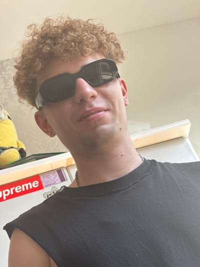 A close-up, slightly upward-angled selfie shows a young person with curly light brown hair and black sunglasses, wearing a black t-shirt. The person's head is tilted slightly to the right. Behind the person, on the left side of the frame, a yellow plush Minion toy is visible on a shelf. To the right of the Minion, a white surface, possibly a refrigerator, is partially visible. On this surface, a red and white "Supreme" logo sticker is prominent. The background features a light-colored wall with a subtle patterned wallpaper. The image appears to be taken indoors in a domestic setting. There is no information in the image to indicate a specific location within the city of Hamm, Germany.