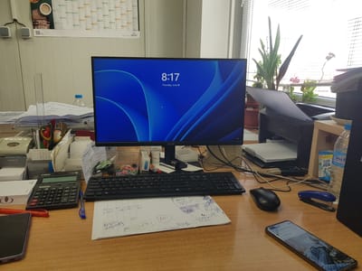 The image depicts a desk with a computer monitor displaying a Windows 11 lock screen with the time "8:17" and the date "Thursday, June 18". A black keyboard and a black mouse are positioned in front of the monitor. To the left of the keyboard, a black calculator with the brand "Canon" is visible. Various office supplies are scattered across the desk, including pens, paper clips, a stapler, a bottle of water, and a smartphone lying face up in the foreground. In the background, a printer is partially visible. The desk is situated in an office environment, with a wall calendar and blinds covering a window. There are no people in the image. The scene is clean and organized, suggesting a workspace. The provided context states the image is located in Lomets, Bulgaria.
