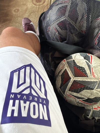 The scene shows a person wearing white shorts with the logo "NOAH YEREVAN est. 2017" in purple, along with a maroon shoe. Next to the person is a soccer ball and a mesh bag filled with other soccer balls. The location is suspected to be the city of Charentsavan, Armenia.