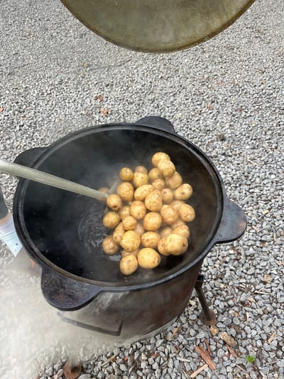 A close-up, high-angle shot shows small, whole potatoes being cooked in a large, black, cast-iron pot. The pot is situated outdoors on a gravel surface, with a large, light-colored lid or cover visible at the top of the frame. Steam rises from the pot, indicating the cooking process. A metal ladle with an ornate handle is being used to stir or remove the potatoes. The surrounding environment consists of grey gravel and a few scattered pieces of wood. The overall scene suggests an outdoor cooking setup. The image is not specific to Sovetskaya Gavan', Russia; it is a generic outdoor scene.