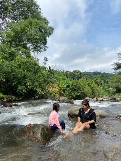 A woman and child sit on rocks in a shallow, rushing stream. The woman wears a dark shirt and shorts, with her legs in the water. The child is positioned on a separate rock, wearing a pink shirt and jeans. The water surrounds them and splashes as it flows. Lush green vegetation covers the riverbank and hillside, extending into the distance under a cloudy sky.