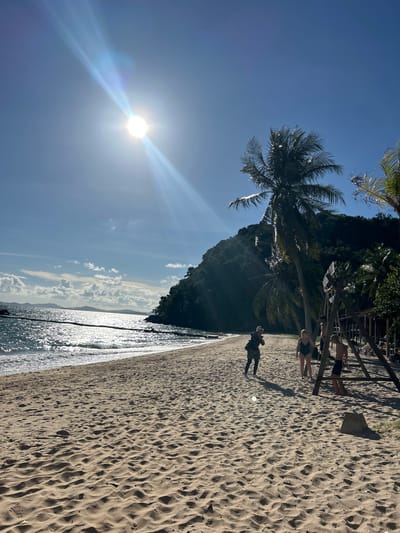 The image depicts a vibrant beach scene in Ban Khlong Hia, Thailand. The sun, shining brightly and creating a lens flare, suggests it's midday. The weather appears to be clear and sunny with a few scattered clouds visible in the distance.

The foreground is dominated by a sandy beach with intricate patterns of footprints. The ocean is a shimmering expanse of blue, reflecting the sunlight. Palm trees with their characteristic fronds are prominent, with one large palm tree leaning over the sand, casting a shadow. In the background, a lush, green, forested hill rises steeply from the shoreline.

Several people are visible on the beach. Closer to the water, a person is walking away from the camera, dressed in darker clothing, possibly walking their dog, which is also visible and appears to be black. Further up the beach, near some wooden structures that might be part of a restaurant or beach bar, a group of people are lounging or standing. Two individuals are clearly visible in swimsuits, one in black and the other in patterned shorts, suggesting a relaxed, holiday atmosphere.

The overall mood of the image is one of tropical tranquility and leisure. The bright sunlight, the calm ocean, and the presence of people enjoying the beach all contribute to a sense of a perfect day in paradise. There is no visible text in the image.