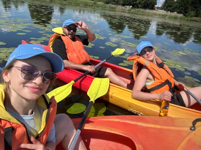 Three people are in kayaks on a body of water, likely a lake or river, surrounded by lily pads and trees. A woman in the foreground is wearing a blue cap that says "STALOGISTIC" and sunglasses, smiling at the camera. She is in a red kayak with an orange life vest. To her right, a man in another kayak is wearing a blue cap, sunglasses, and an orange life vest. He is looking at the camera and appears to be talking. Behind him, a woman in a yellow kayak is wearing sunglasses and an orange life vest. She is holding a bottle of drink. Two yellow kayak paddles are visible. The water reflects the sky and trees. The scene appears to be in a rural area. The presence of "STALOGISTIC" on the caps suggests a possible connection to the company Stalogiistic located in the village of Basaliowszczyzna, within the Valozhyn District, Minsk Region, Belarus.
