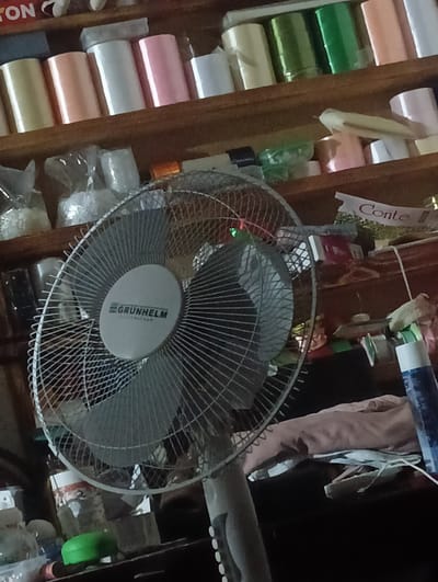 The image shows a close-up of a standing fan with a metallic grille and a central circular logo that reads "GRUNHELM". The fan is positioned in front of shelves filled with various items, including rolls of colorful ribbon or fabric, plastic-wrapped objects, and what appear to be boxes. In the background, a white banner with red lettering, possibly a logo or advertisement, is visible, reading "Conte". The overall impression is of a cluttered storage area or shop. The image is taken indoors, and there are no people visible. The setting does not provide any specific clues about its location in Bender, Moldova.