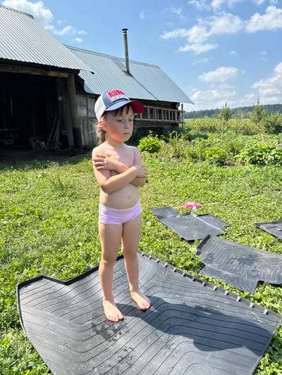 A young girl, wearing a cap and pink underwear, stands with her arms crossed in a grassy yard. She is positioned in front of a rustic building with a corrugated metal roof and a chimney. The background features lush green foliage and trees, suggesting an outdoor, rural setting. In the foreground, several black rubber floor mats are laid out on the grass. The sky is visible above with scattered clouds, indicating it is daytime, likely a sunny day. The overall impression is one of a casual, everyday moment in a summery environment. The cap the girl is wearing has text and a logo on it.
