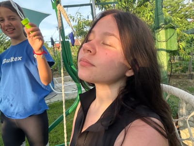 The image depicts an outdoor scene in what appears to be a residential garden or yard in Byala, Bulgaria, illuminated by warm, direct sunlight suggesting late afternoon or early evening.

On the left, a young female, likely a pre-teen or early teen, is partially visible, smiling broadly towards the viewer. She has dark brown hair and wears a blue short-sleeved t-shirt with "Bultex" printed in white across the chest, and black leggings that appear to have a white lace detail near the ankles. Her right arm is raised, holding a neon green collapsible bubble wand. A red braided string bracelet with a silver heart charm and a darker, possibly animal-shaped, charm is visible on her right wrist.

To the right, another young female, also with long dark brown hair, is seated with her eyes closed, face tilted upwards and slightly to the left, directly exposed to the sunlight. She wears a black sleeveless top with a prominent vertical zipper down the front. Her expression is serene and relaxed, seemingly enjoying the warmth of the sun. Minor skin redness or blemishes are visible on her cheeks and chin. She is positioned within what appears to be a swing or hanging chair made of white braided rope, partially visible on the right side of the frame.

The immediate background features lush green foliage, including trees and grass covering the ground. A weathered, bright green painted metal pole or frame, possibly part of a swing set or garden structure, stands vertically between the two individuals. In the more distant background, a clothesline with various garments (a blue shirt and a darker item) is visible hanging between unseen points, against a backdrop of more greenery and hints of distant residential structures with light-colored roofs. White pipes or hoses are also visible on the ground near the left subject. The sky is a clear, light blue.