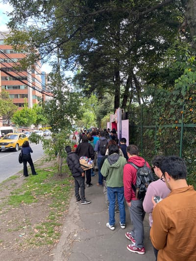 This image captures an outdoor urban scene in Bogota, Capital District, Colombia, during daytime under a partly cloudy sky.

A prominent feature is a long queue of approximately 30-40 individuals, predominantly adults, standing in a single file on a cracked concrete sidewalk. The queue extends from the foreground towards the background, moving along the right side of the frame. Most individuals are facing forward, dressed in casual clothing including jackets, hoodies, and jeans. One male individual in the mid-ground, positioned slightly to the left of the queue, carries a wooden tray filled with various packaged items, indicative of a street vendor. Another female individual with dark hair is visible on the far left of the sidewalk, near the street, appearing to observe the scene.

To the immediate right of the queue, a green metal fence is covered by dense green vines interspersed with orange flowers. This fence borders a heavily vegetated area dominated by large, mature trees that provide a dense canopy overhead.

On the left side of the image, an asphalt road with at least two lanes is visible. A yellow taxi, characteristic of Bogota, is prominently positioned in the left lane, moving away from the viewer. Other vehicles, including a white van and cars, are discernible further down the street. Multi-story buildings with brick or concrete facades, featuring numerous windows and balconies, form the urban backdrop beyond the road. Overhead utility wires are visible traversing the scene between these structures.

At the perceived end of the queue, in the distant right, two light-colored, temporary structures or banners are partially visible. One displays the word "LOVE" with a heart symbol, and another exhibits the numerical sequence "1-1-8" along with pink elements, suggesting an organized event or designated entrance. The unpaved ground adjacent to the sidewalk shows sparse vegetation.