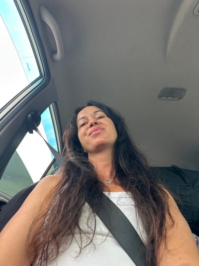 The image shows a woman sitting in the passenger seat of a car. She is looking up and to her right. She has long, dark hair and is wearing a white tank top and a necklace. A black seatbelt is visible across her chest. The interior of the car is visible, including the headliner and a grab handle. The sky is visible through the car window. The location is the city of Slyudyanka, Russia.