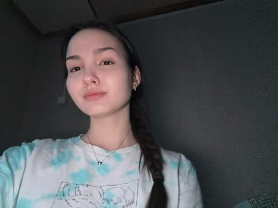 A single female individual is depicted in an indoor setting within the city of Bayronovka, Russia. The subject, a young adult with fair skin and dark brown hair styled in a braid over her left shoulder, is positioned facing the camera with a neutral expression and a slight closed-lip smile. She wears small silver stud earrings in both ears, with an additional hoop or cuff in her right ear. A silver-colored chain necklace with a small, circular, dark-colored pendant is visible around her neck. Her attire consists of a short-sleeved t-shirt featuring a white base with teal tie-dye patterns and a black-and-white graphic print depicting a landscape. The background behind the subject is a dark, textured wall, possibly charcoal grey, with a darker horizontal beam or frame visible at the top of the image. The lighting is dim and appears to emanate from the front-left, illuminating the subject's face.