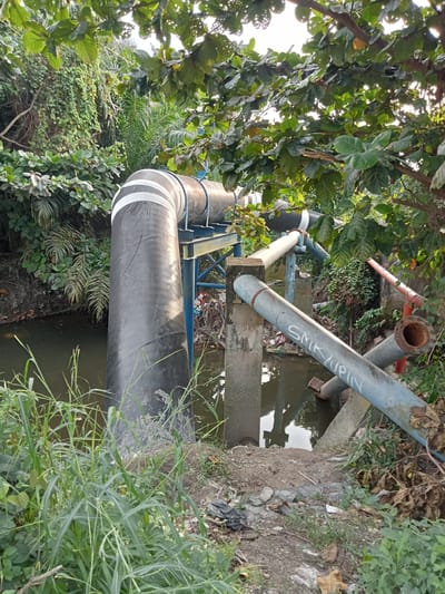 The image depicts a large, industrial pipe system partially submerged in a canal or drainage ditch in Kubang Jaya, Indonesia. The main pipe, which is black with white bands, is supported by a blue metal structure. Several smaller, lighter colored pipes are also visible, some with visible writing that appears to read "SMK JUPIN". These pipes are supported by concrete pillars that are partially in the water. The surrounding environment is overgrown with vegetation, including tall grasses, vines, and leafy trees. Debris, including plastic bags and what appears to be trash, is visible along the banks of the canal and in the water. The scene suggests an industrial or utility area adjacent to a natural or semi-natural waterway. The lighting suggests it is daytime, possibly late afternoon due to the direction of the light. No people are visible in the image.