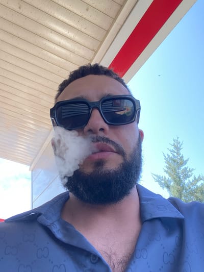 A low-angle, medium shot captures a man exhaling smoke while looking up at the sky. He is wearing black sunglasses and a dark blue shirt with a repeating Gucci logo pattern. Smoke emanates from his mouth and obscures part of his face. The background features a wooden ceiling and a clear blue sky with a thin tree on the right. The image appears to have been taken outdoors, possibly at a gas station, given the structure of the ceiling and the bright lighting. The specific location in Korkmaskala, Russia, is not discernible from the visual cues in the image.