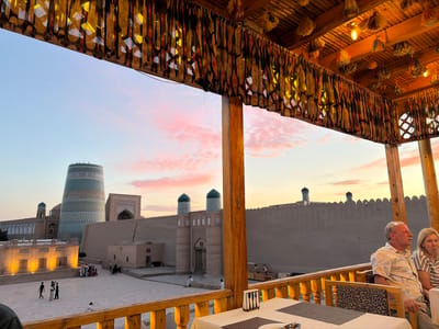 The image depicts an evening scene from a raised, covered outdoor seating area overlooking a historical courtyard in Khiva, Uzbekistan. In the foreground, a wooden railing with decorative carvings frames the view. A table set for dining is visible, with a white tablecloth, placemats, and cutlery. Two individuals, a man and a woman, are seated in chairs facing away from the camera, looking out at the scene.

The courtyard below features ancient architecture characteristic of Khiva. Dominating the left side of the frame is the iconic Kalta Minor minaret, a truncated turquoise tower adorned with intricate blue and white patterns. Adjacent to it are other historical buildings, including a mosque with green-domed minarets and a long, imposing wall constructed of mud bricks. Several people are scattered throughout the courtyard, some walking and others gathered near stalls, suggesting a marketplace or tourist area. The sky is a soft gradient of pink and orange, indicating sunset. The covered area where the photo is taken has a wooden roof adorned with decorative fabric hangings and dangling tassels, illuminated by warm lighting.