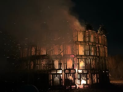 A massive bonfire built from wooden pallets burns intensely against a dark night sky. Flames are visible through the pallet structure, and smoke billows upwards. Sparks fly off the fire, adding to the hazy atmosphere. The bonfire appears to be several stories tall and sits on a patch of dark ground. Small trees are positioned strategically on the upper levels.