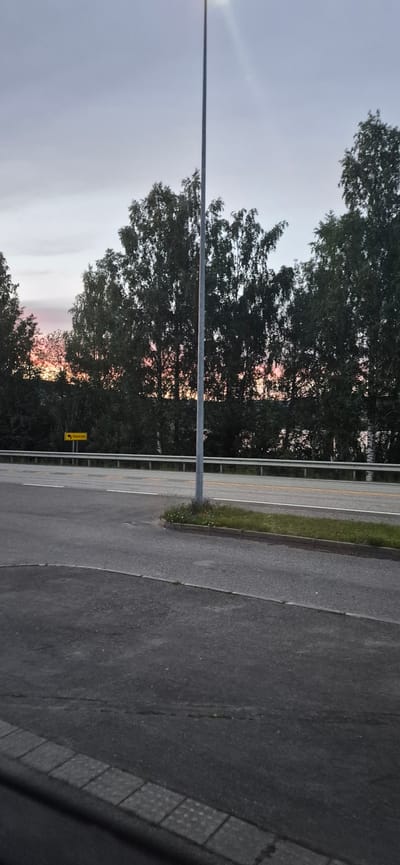 This media file shows a view from a roadside in Biri, Norway, during what appears to be either sunrise or sunset. The sky is a soft gradient of pale blue and pink hues, suggesting either the early morning or late evening. Tall, dark green trees line the left side of the image, their dense foliage partially obscuring the colorful sky. A tall, grey utility pole stands prominently in the foreground, running vertically through the frame.

To the right of the trees, a paved road is visible, with distinct lanes marked by yellow and white lines. A strip of green grass and small wildflowers separates the road from a metal guardrail that runs along its edge. The road itself is asphalt, showing signs of wear. Further to the right, the surface texture changes to a darker, more textured material, possibly a different section of the road or a parking area.

A small, yellow rectangular sign with a black arrow pointing to the left is attached to the guardrail, indicating a direction. The text on the sign appears to be "Hanssya". The overall mood is peaceful and tranquil, with the natural beauty of the landscape highlighted by the soft lighting. The time of day is suggested by the gentle, diffused light and the colors in the sky. There are no people visible in the frame.