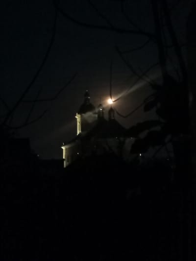 An image of a building with multiple domes is visible against a dark night sky, illuminated by the moon which is partially obscured by clouds and branches. The building appears to be a church or a similar religious structure, possibly located in Yampil, Ukraine, as suggested by the context. The moon's light casts a beam through the clouds, creating a dramatic effect. Silhouetted branches and leaves from nearby trees are in the foreground, obscuring parts of the view and adding depth to the scene. The overall impression is one of a quiet, moonlit night with a prominent architectural feature in the background. There are no people or visible signs of activity in the image.