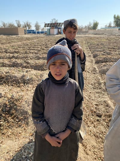 The image portrays two young males and a partial third individual standing in a tilled field under a clear blue sky in Mandisar, Afghanistan. In the immediate foreground, a child, estimated to be between 6 and 9 years old, faces directly forward, establishing eye contact with the viewer. This child wears a blue and grey knitted beanie with an orange stripe, a dark grey turtleneck, and a dark tunic adorned with a lighter grey, patterned sleeveless overlay on the chest. Their hands are clasped in front of the waist. Positioned behind and to the right is an older male, appearing approximately 10 to 14 years old, who wears a light grey, open-weave cap and dark garments. This individual holds a wooden-handled shovel or hoe vertically, with the blade resting on the ground, and looks slightly to the right of the frame. On the far right edge, a partial view of a light-colored garment belonging to a third person is visible. The ground consists of dry, light-brown soil arranged in distinct furrows, indicative of agricultural activity. The background features several structures: a light-brown mud-brick building, a compound with blue fencing, and a prominent solar panel array positioned on a framework. Sparse, bare trees are scattered in the mid-ground, beneath a cloudless blue sky.