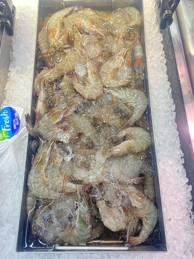 The image presents an overhead view of a refrigerated display case filled with fresh, raw prawns (shrimp) and crushed ice. The setting appears to be a seafood counter in a supermarket or fish market, located in Moo 1, Thailand.

Hundreds of whole, shell-on prawns, predominantly light greyish-green in color, are tightly packed within a rectangular metal container. They are largely submerged in water, which, along with the generous amount of crushed ice, serves to keep them chilled and fresh for sale. Some prawns display delicate reddish antennae or legs. A pair of black metal tongs is visible resting on the ice in the upper left corner, suggesting a self-service arrangement for customers to select their seafood.

A blue and white plastic label or bag is partially visible at the bottom of the frame. The text "fresh" is prominently displayed in blue stylized lettering, with "SPECIAL SELECTED" visible above it, printed upside down. Part of "We care" is also discernible. The lighting is bright and uniform, characteristic of an indoor retail environment. No individuals are visible, keeping the focus entirely on the seafood display.