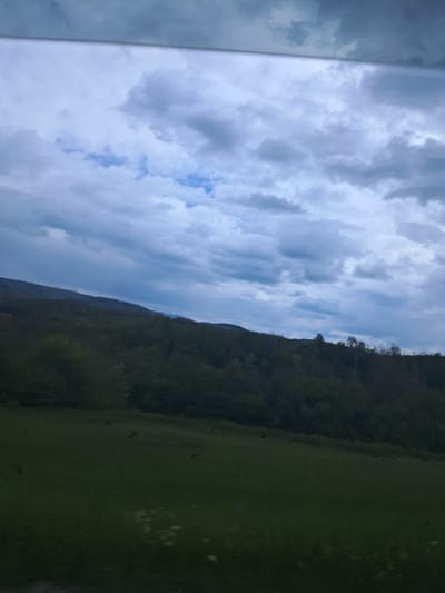 The image shows a view of a cloudy sky and a landscape, possibly taken from inside a building or vehicle, as indicated by the reflective surface on the left edge of the frame. A significant portion of the image is occupied by an overcast sky filled with grey and white clouds. Below the sky, a hill or mountain range covered in dense forest is visible. A green field extends further to the right. The exact location is suggested to be in the city of Boltata, Bulgaria.