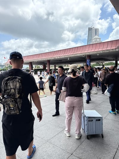 In Xiangzhou, China, an outdoor scene shows a crowd of people, some walking and others standing. In the foreground, a person wearing a camouflage backpack and black t-shirt walks away from the camera, while another person in a light-colored hat and pink pants is seen from behind, pulling a light blue folding cart. Behind them, a group of people, including men and women of various ages, are gathered under a covered walkway with a red-tiled roof. Some individuals carry bags, and one person is holding a shopping bag. In the background, a tall, modern building with a distinctive pointed roof stands against a cloudy sky. The ground is paved with square tiles.