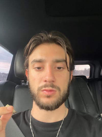 A man is sitting in the driver's seat of a car. He is holding a rolled cigarette in his left hand, which is positioned near his mouth. He is wearing a black t-shirt and a silver chain. The car appears to be a luxury SUV, with black leather seats and a sunroof. The exterior view through the window suggests a suburban or semi-rural environment, possibly in the late afternoon or early evening, with a pale sky and distant buildings or hills. The setting is consistent with a location in Ciudad López Mateos, Mexico, a municipality known for its mix of urban and suburban areas.