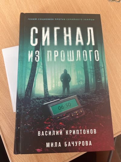 The image shows the cover of a book titled "СИГНАЛ ИЗ ПРОШЛОГО" (Signal from the Past). The cover depicts a dark, foggy forest scene with a silhouetted figure wearing a hooded jacket standing in the center. In the foreground, a pager-like device is visible with the display showing "06:50". The book cover also features text that reads "ГЕНИЙ-СОЦИОФОБ ПРОТИВ СЕРИЙНОГО УБИЙЦЫ" (Genius-Sociophobe vs. Serial Killer) at the top, and author names "ВАСИЛИЙ КРИПТОНОВ" (Vasily Kryptonov) and "МИЛА БАЧУРОВА" (Mila Bachurova) at the bottom. The book is placed on a wooden desk. There is no information about the location in the city of Toila, Estonia, in this image.
