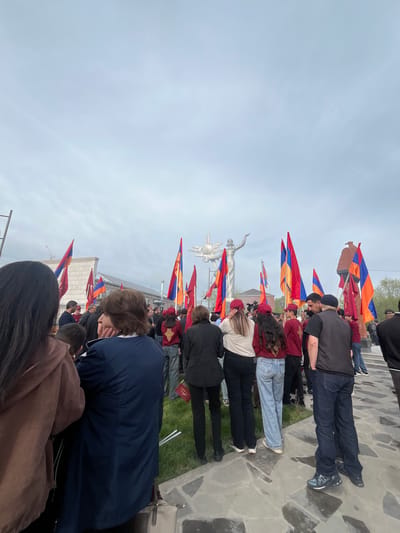 The image shows a group of people in a public space, potentially a memorial or gathering site, in Akhuryan, Armenia. They are holding Armenian flags, identifiable by the horizontal tricolor of red, blue, and orange. In the background, there's a statue of a woman holding a sword, likely a monument. The crowd is gathered on a paved area, possibly a plaza, with some grass visible between the stones. People are facing the statue. The sky is overcast. Some of the people have their heads bowed. The scene is outdoors, and it seems like a public event or a memorial gathering.