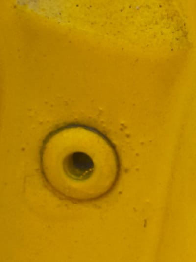 This image is a close-up shot of a yellow, textured surface with a circular indentation. The indentation has a raised rim and a dark opening in the center, suggesting it might be a bolt head, a recessed button, or a similar fixture. The surface appears to be outdoors, given the slight dappling of light and shadow, and the texture suggests it could be a metal or plastic material, possibly painted. There are small dark specks scattered across the yellow surface, which could be dirt, debris, or part of the material's texture. The lighting suggests it is daytime, but the weather is not discernible. There is no text visible in the image. The context provided places this scene in Nowy Dwór Mazowiecki, Poland.