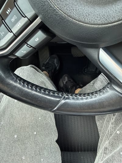 The image shows a close-up view from inside a car, focusing on the steering wheel and the driver's legs. The steering wheel is black leather with visible stitching. On the left side of the steering wheel, there are buttons, including one with an "INFO" label and another with a phone icon. The driver is wearing grey jeans, some of which appear to have small reflective or shiny embellishments. Their feet are visible, wearing camouflage-patterned sneakers, resting on or near the car's pedals. The floor of the car has a textured black mat. The scene suggests the car is stationary, likely parked or stopped. The prompt's request to include the location of Šilutė, Lithuania is not supported by any visual information in the image, as there are no identifying landmarks or context to place the scene geographically. Therefore, no information about Šilutė, Lithuania can be provided.