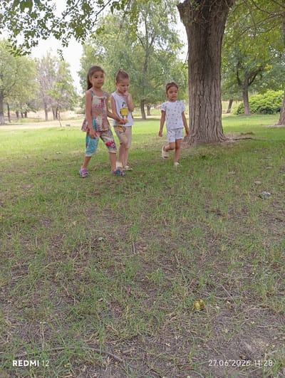 Three young children are standing on a grassy area in a park-like setting. The child on the left is a girl wearing a patterned sleeveless top and patterned pants. The child in the middle is a boy wearing a white tank top and light brown shorts. The child on the right is a girl wearing a white t-shirt with a printed pattern and white shorts. They are all looking in different directions, and the boy has his finger to his mouth. Behind them are several trees, and the ground is covered with grass and dry leaves. The overall impression is a casual outdoor scene. The date and time stamp at the bottom right of the image indicates "27.06.2026 11:28".