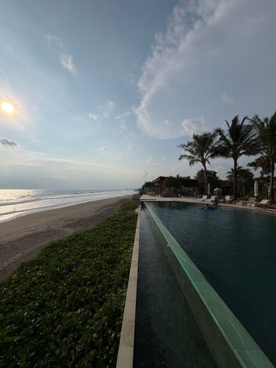 The image displays a coastal scene in Belalang, Indonesia, captured during late afternoon or early morning. A wide expanse of dark grey sand beach extends from the left midground towards the right, meeting the dark greyish-blue ocean where white waves break repeatedly. The sun, a bright yellow sphere, is low on the horizon in the upper left, casting a strong glare across the ocean's surface and illuminating the underside of scattered white clouds in the light blue sky.
Running parallel to the beach, a dense strip of low-lying green groundcover separates the sand from a resort or hotel complex. The foreground is dominated by a dark-water infinity-edge swimming pool, its greenish-blue textured wall forming a waterfall effect into a lower trough. One individual, partially submerged, floats in the pool on a blue inflatable ring, facing towards the right.
Beyond the pool, a paved area features multiple white lounge chairs, some with striped cushions, and several large, dark beanbag-style chairs. Two individuals are seated on these chairs near the pool's edge, partially obscured by open white beach umbrellas and tropical foliage. Further back, numerous tall palm trees line the property, and several resort buildings are visible, including a prominent open-air structure with a dark roof and wooden pillars. A small, white conical cabana or tent is also present in the lounge area.