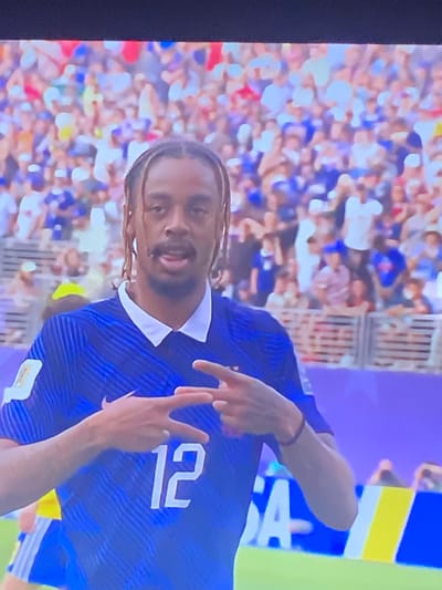 The image shows a male soccer player wearing a blue jersey with white stripes and the number 12 on the front. He has dreadlocks and is looking at the camera with his mouth slightly open. His hands are raised, and he is forming a gesture with his fingers. The player is standing on a soccer field with a blurred crowd of spectators in the background. The crowd is wearing a variety of colored clothing, predominantly blue and white. There are also some yellow and red colors visible in the crowd. The background also shows parts of a stadium with railings and banners. The lighting suggests it is daytime. The image does not provide any specific details to indicate a location within the city of Zastavna, Ukraine.