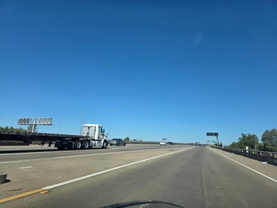 A wide-angle shot captures a bright, clear day on a multi-lane highway, likely in Sacramento, USA. The sky is a vibrant, cloudless blue, suggesting good weather. The road is asphalt, with distinct white lane markings and a yellow line on the left shoulder.
In the foreground, a flatbed truck with a white cab and a long, empty trailer is traveling in the right lane. The truck has multiple axles and large rear wheels. Further down the highway, several other vehicles are visible, including a silver sedan, a white car, and some cars in the distance. The scene appears to be during the daytime, with the sun casting shadows and illuminating the road.
To the left of the highway, a large billboard structure is visible, supported by a tall metal frame. On the right side of the road, trees line the median and the edge of the highway. The overall atmosphere is one of travel and transportation on a typical sunny day. There is no visible text in the image.