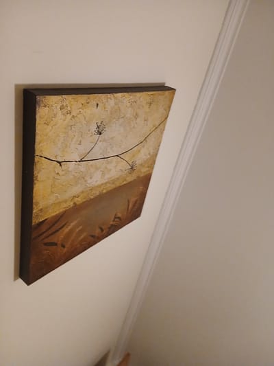 A framed canvas painting featuring a minimalist depiction of dried plants is mounted on an off-white wall. The painting is hung at an angle, showing a dark brown frame around a textured canvas. The upper portion of the canvas is a light, textured beige with subtle yellow undertones, while the lower portion transitions to a darker brown with abstract, sweeping brushstrokes that suggest reeds or grass. A thin, dark line extends diagonally across the upper half of the canvas, with two small, delicate dried seed heads attached to it. To the right of the painting, a white architectural molding with a fluted design runs vertically up the wall. The scene is illuminated by warm, artificial light, casting soft shadows. The background is a plain, off-white wall. The painting appears to be in a domestic setting, possibly a living room or hallway, in Póvoa de Varzim, Portugal.