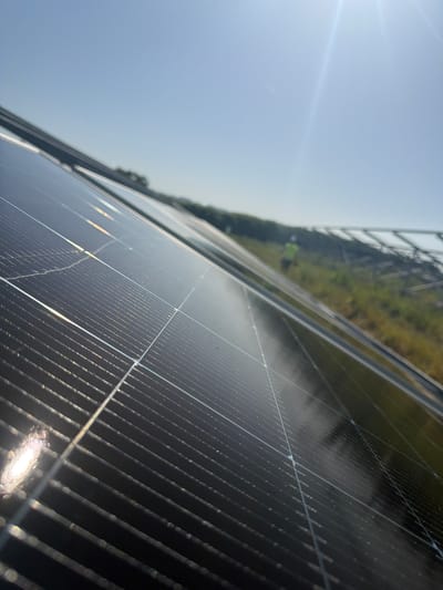 The image shows an outdoor scene with a close-up perspective of solar panels. The solar panels are primarily black with metallic lines visible on the surface. There is a glare from the sun on the panels, suggesting a sunny day. In the background, there's a glimpse of greenery and some infrastructure that could be part of the solar panel installation. The location is Lacock, United Kingdom.
