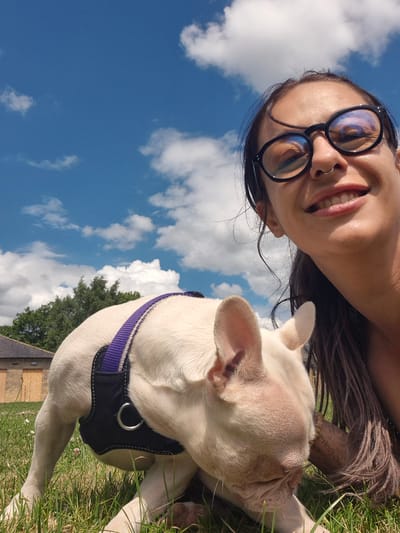 This media file is a selfie featuring a woman and a dog outdoors. The woman is wearing glasses and smiling at the camera. Her dark hair is visible, and she appears to have a nose ring. A white French bulldog, wearing a black and purple harness, is nuzzling against her face. They are in a grassy area, with a building and trees in the background. The sky above is bright blue with fluffy white clouds, suggesting it is daytime and the weather is fair. The image is oriented sideways, with the subjects at the bottom and the sky at the top. There is no visible text in the image. The location context is Bridlington, United Kingdom. The overall emotion conveyed is one of happiness and companionship between the woman and her dog.