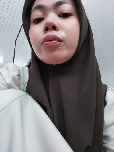 This is a close-up, selfie-style, horizontal, eye-level shot of a young woman wearing a brown hijab and light-colored clothing. The woman is looking directly at the camera, with her face slightly angled. Her makeup includes defined eyebrows, eyeliner, mascara, and glossy pink lipstick. The background consists of a white ceiling with visible parallel lines, suggesting an indoor setting, possibly a room with a slatted ceiling or ventilation system. The lighting appears to be artificial and soft, indicating it could be daytime or nighttime. The overall mood is casual and personal, typical of a selfie. The image does not contain any visible text. The provided location context of Tibojong, Indonesia is external information and not derivable from the image itself.