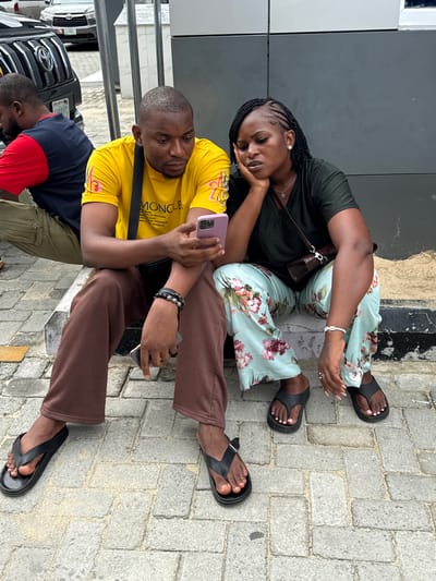 Two individuals are seated on the ground in an urban setting in Ojomu, Nigeria. The woman, who is on the left, wears a dark green top and floral print pants, with her head resting on her hand. The man wears a yellow shirt and brown pants and is holding a mobile phone. Both are wearing sandals. The background shows a dark gray wall and a brick pavement. A car is partially visible in the lower left corner.