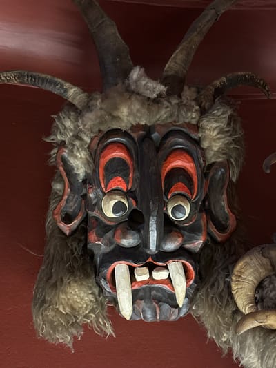 The image shows a close-up of a Krampus mask, a traditional figure associated with Austrian Christmas customs. The mask is made of dark wood with bright red and black accents. It features large, bulging eyes with red and black surrounds, and pronounced white teeth, including two long, sharp fangs. The mask is adorned with fur and has prominent, curled horns made of real animal horns. One ram's horn is visible to the right of the mask. The background is a plain, dark red surface, suggesting it is likely displayed indoors, possibly in a museum or cultural exhibit. While the image itself does not explicitly show the city of Salzburg, Austria, Krampus traditions are particularly strong in the Alpine regions of Austria, including Salzburg. The mask is a cultural artifact, likely used in folk performances or as a decorative item.