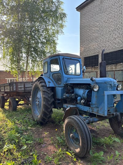 The image depicts an old blue tractor hitched to a wooden trailer, parked on a grassy area next to a brick building. The tractor is positioned in the foreground, facing right, with its front wheel prominently displayed. Its engine is partially exposed, revealing various mechanical components. The trailer behind it appears worn and weathered.

The background features a light-colored brick building with horizontal window-like openings covered by dark louvers. Tall trees with green foliage, possibly birch trees, are visible to the left of the tractor, with their white bark contrasting with the greenery. The sky is clear and blue, suggesting it is daytime.

The scene is likely an outdoor area, possibly a farm, industrial yard, or a storage area for agricultural equipment in the village of Bolshiye Usy, Russia. There are no people present in the image. The overall impression is one of a functional but aged piece of machinery situated in a somewhat rural or semi-industrial environment.
