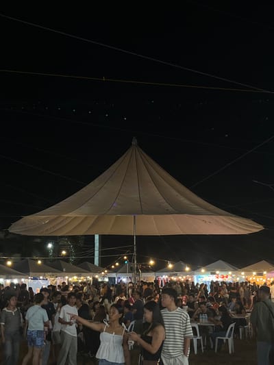 The image depicts an outdoor night market event, likely a festival or bazaar, in Koronadal City, Philippines. A large, light-colored tent-like structure with a conical peak dominates the center of the frame, serving as a central canopy. Numerous smaller tents, typical of market stalls, are visible in the background, illuminated by strings of yellow lights, creating a festive atmosphere. The ground appears to be a dirt or grass area, with many people milling about.
The foreground shows a crowd of individuals, with varying degrees of focus. Several people are seen in the mid-ground, some sitting at tables, suggesting food vendors or dining areas. The overall scene is characterized by a lively gathering under the night sky, with the characteristic infrastructure of a temporary market. The presence of numerous people, stalls, and lighting strongly indicates a public event occurring after sunset.