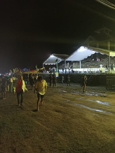 This night-time image captures an outdoor public event in Kecamatan Pujut, Indonesia. The scene is illuminated by artificial lights, contrasting with the dark sky above.
In the foreground, two individuals walk towards the viewer on an uneven dirt path, which appears wet or muddy in places, reflecting light. One person wears a bright yellow short-sleeved shirt and dark shorts, while the other is dressed in a red shirt and a light-colored jacket. Numerous other people are scattered across the middle ground, some standing and observing. Several individuals in uniform, possibly security personnel, are visible near the temporary structures.
The background features two prominent white canopies, brightly lit from beneath, suggesting stages or viewing areas. White chairs are arranged in rows in front of these canopies, indicating audience seating. Behind these structures, an elevated, terraced area, possibly made of stone or concrete, holds more attendees. To the far left, several smaller tents and numerous flags add to the festive atmosphere. A taller, multi-story building or structure is partially visible behind the canopies to the right.
The overall impression is that of a bustling outdoor gathering or festival taking place at night, with temporary setups and a significant number of participants and observers. The damp ground suggests recent rain or general moisture in the area. There is no discernible text visible in the image.