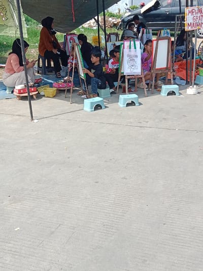 The scene takes place in Kendal, Indonesia, at the coordinates -6.932 latitude and 110.203 longitude. A group of people are gathered under a canopy, presumably for an art lesson. There are at least seven children, some sitting on small stools and drawing on easels. Three adults, a man and two women, appear to be supervising the lesson. A woman in a hijab is seated to the left of the image, looking at a mobile phone, while another woman in a hijab sits to the right of the image, watching the children. The children are drawing on whiteboards mounted on easels. There is a sign to the right of the image, but the text is not legible. The ground is a light gray concrete, with multiple linear grooves. There is a lot of clutter in the background, but it is difficult to discern.